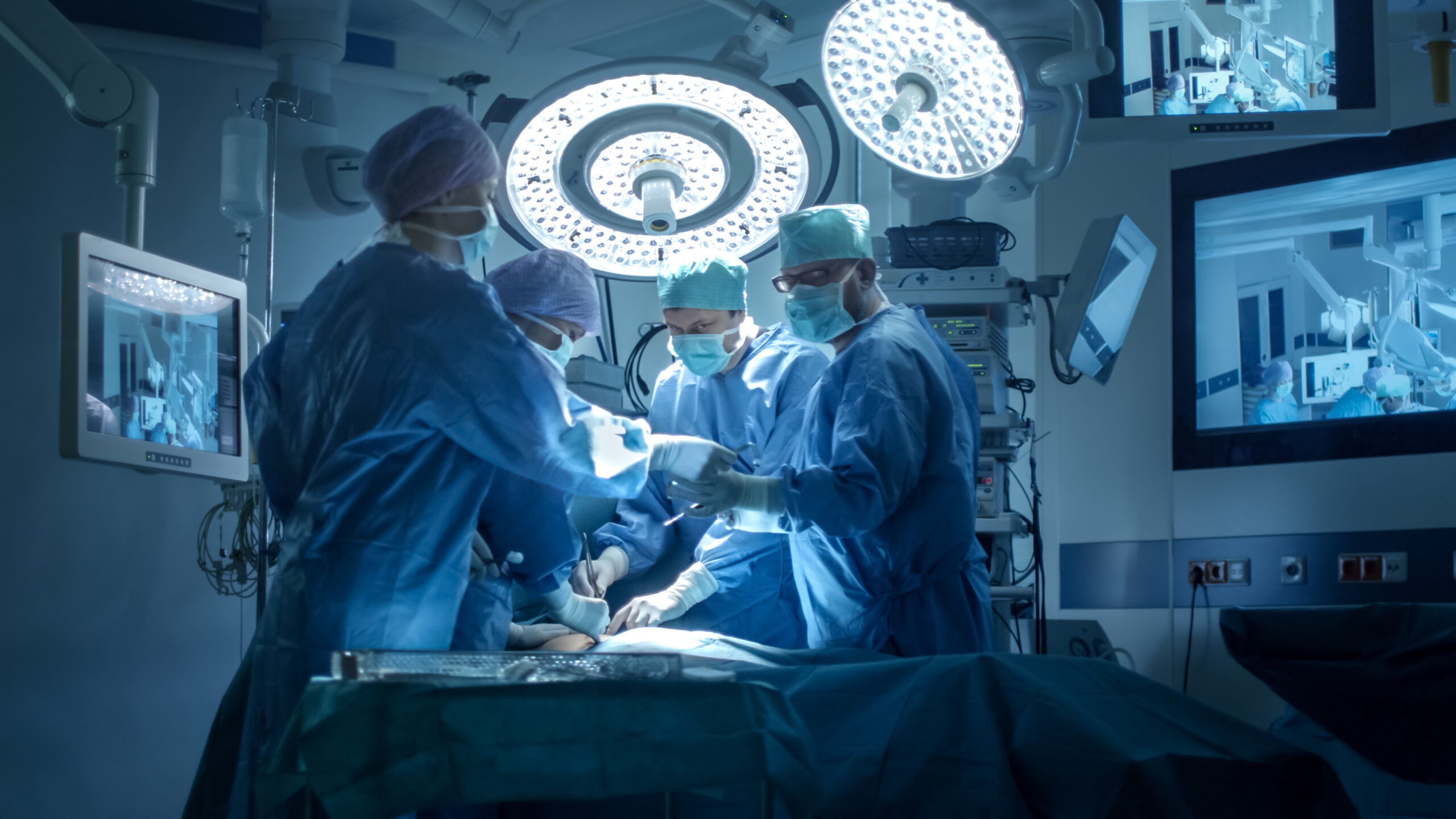 Four surgeons in an operating room with surgical screens in the background.